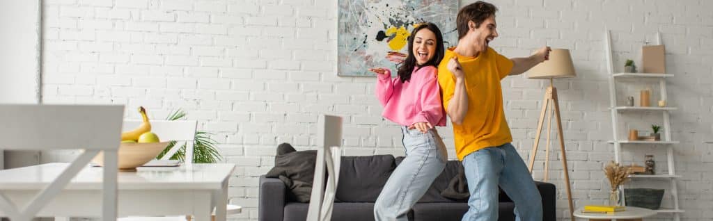 smiling young couple in casual clothes dancing in living room banner